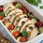 Mediterranean chicken zucchini bake 3 A flat-lay shot of all the ingredients for the Mediterranean Chicken Zucchini Bake laid out on a clean white surface. The ingredients include boneless chicken breasts, sliced zucchini, a bowl of cherry tomatoes, a jar of olives, a block of feta, and fresh herbs.